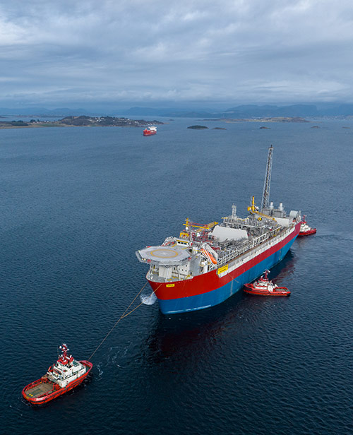 Aerial view of the Jotun FPSO, a large red and blue floating production storage and offloading vessel, being towed by tugboats across the open sea.