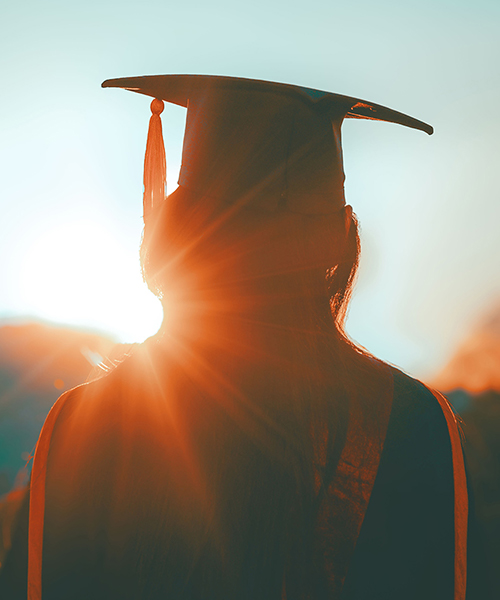 Person wearing graduation gown and cap in front of a bright sun.