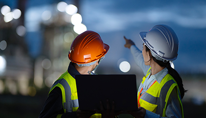 Two people wearing hard hats looking over towards an illuminated refinery.