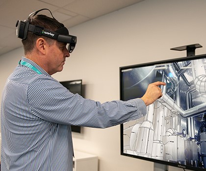 Person wearing a virtual reality headset looking at and pointing to a digital twin on a TV screen.